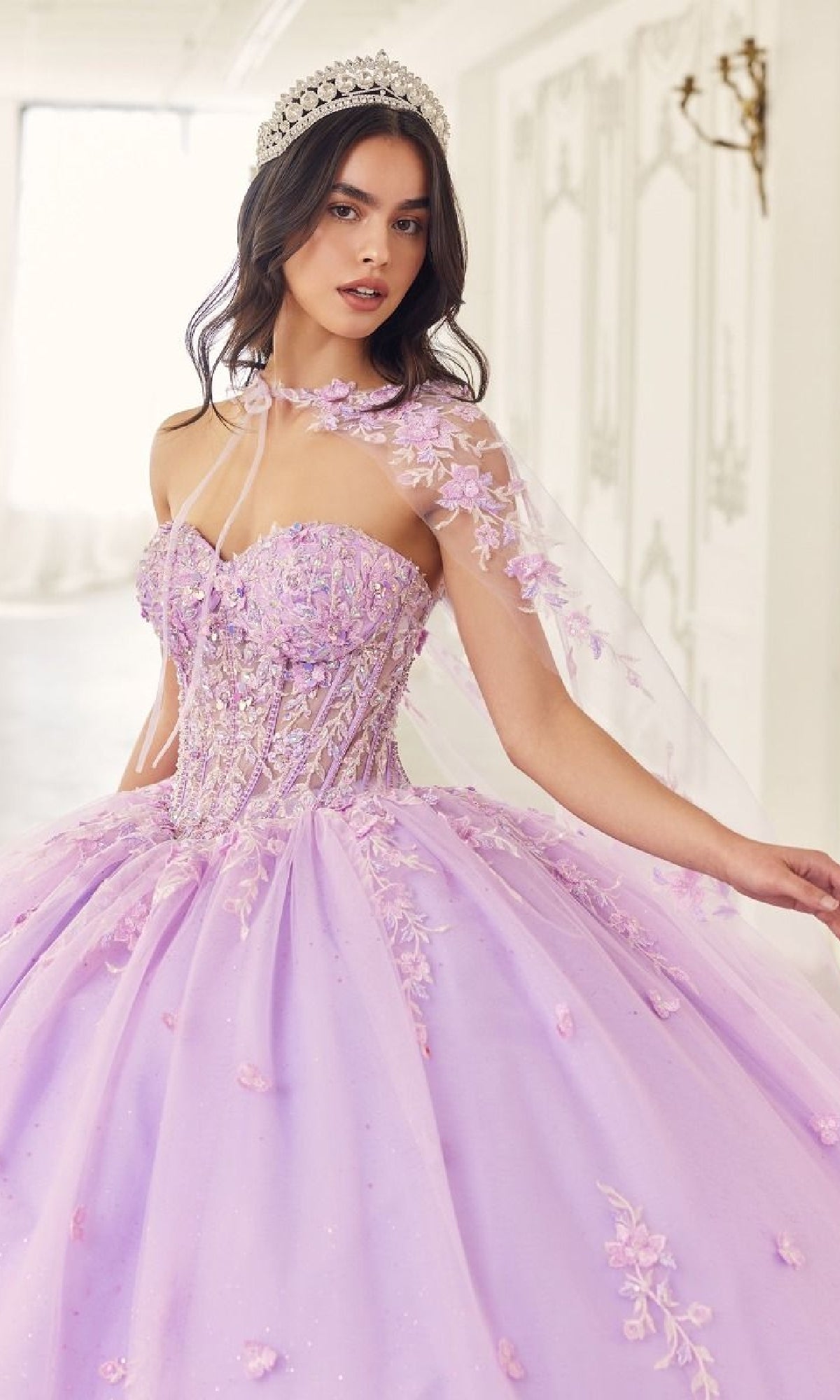 A young woman stands in a light-filled room wearing the Quinceanera Dress 15723 by Ladivine—a lavender ball gown with a corset bodice, floral embroidery, sheer cape, and jeweled tiara.